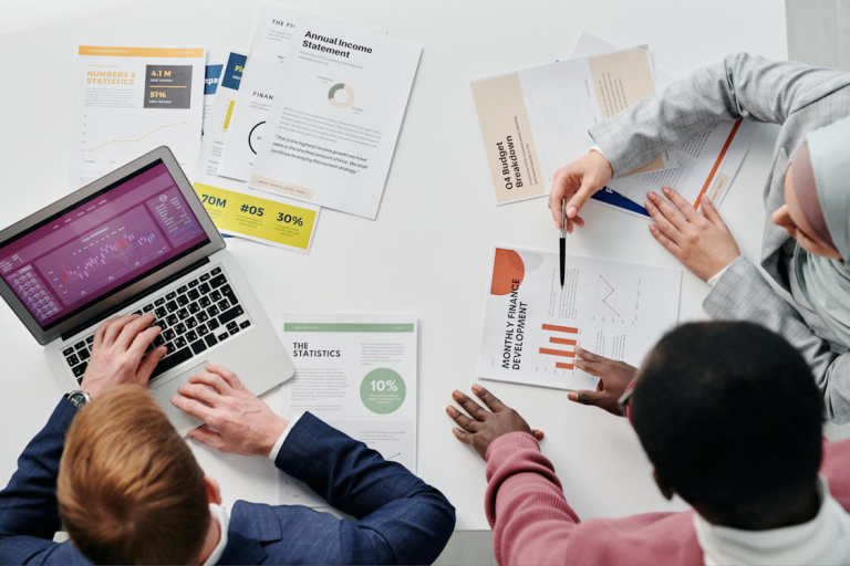 Three people sitting at a desk reviewing business documents, including an annual income statement and statistical reports.