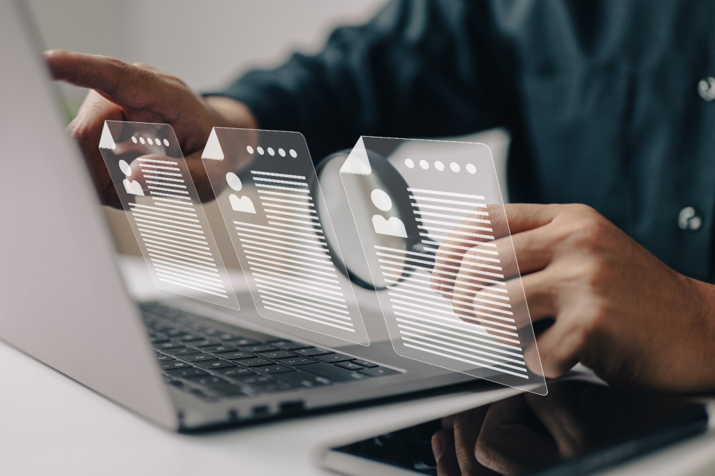 An image shows a person holding a magnifying glass using a laptop with a phone beside it and a graphic of three CVs representing the benefits of HRMS for businesses looking to streamline candidate applications and facilitate resume screening.