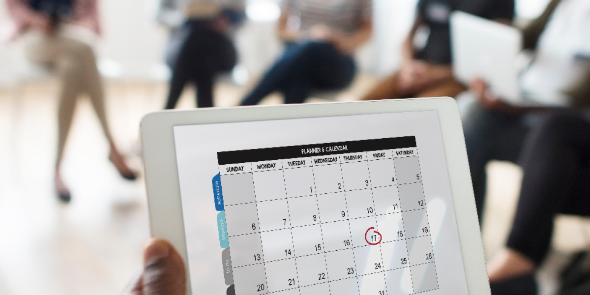 A person holding a tablet displaying a scheduled calendar while a team collaborates in the background.