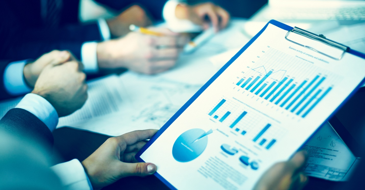 Hands are seen holding a blue clipboard with a printout of data in various pie charts and graphs, while other hands are seen in the background going over paperwork.