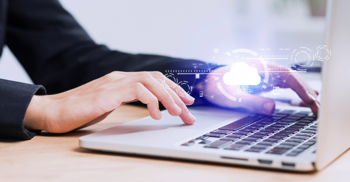 A person’s hand is seen on the touchpad of an open laptop with the other on the keyboard, while a graphic shows a white cloud to highlight the importance of cloud security for businesses leveraging cloud computing and storage.