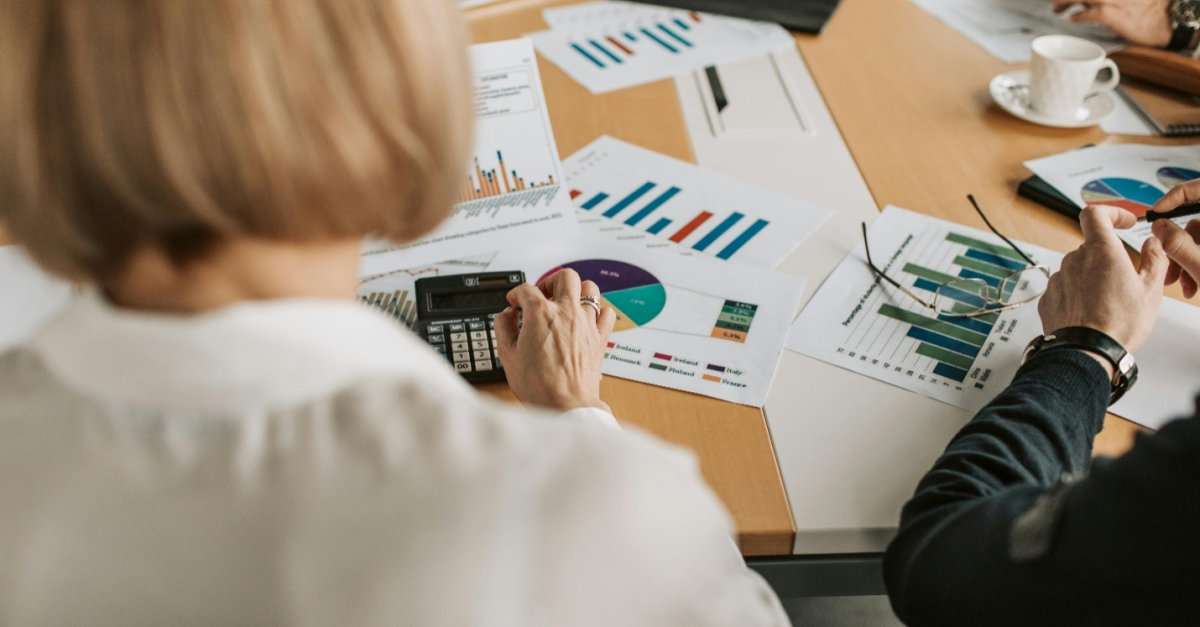 Desk scattered with papers showing data on graphs and charts and stationery, surrounded by a couple of people, highlighting how Sage 300 or Sage Intacct are a great choice for data analysis and management.